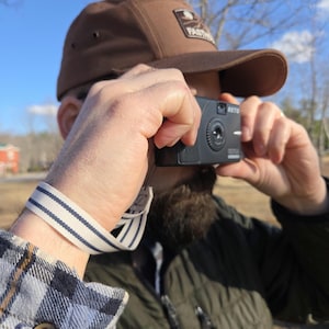 Könnte beinhalten: Eine schwarze RETO-Kamera wird von einer Person gehalten, die eine braune Kappe mit einem Logo trägt. Die Person trägt auch ein blau-weiß kariertes Hemd und eine grüne Jacke. Die Kamera hat ein Objektiv und das Wort RETO ist darauf gedruckt.