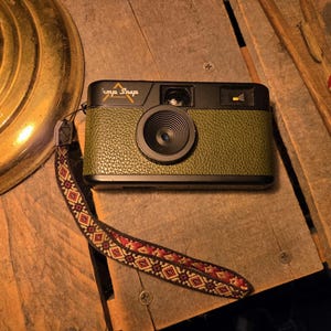 May include: A black and olive green camera with the text "imp Snap" on top. The camera has a textured leather-like grip and a patterned strap. The camera is on a wooden surface.