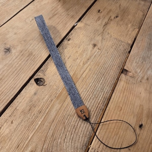 May include: A blue denim wrist strap featuring a tan leather connector tab with a small black geometric logo embossed on the surface. The strap includes a thin black cord loop at the end for attachment to small electronic devices or accessories.