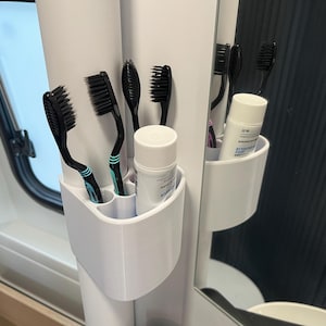 Puede incluir: Un soporte blanco para cepillos de dientes con varios cepillos de dientes negros y un tubo de pasta de dientes. Los cepillos de dientes tienen cerdas negras y algunos tienen detalles turquesa. El tubo de pasta de dientes es blanco con texto azul.