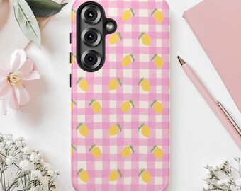 May include: A pink and white checkered phone case featuring a pattern of yellow lemons with green leaves. The case has a black camera module and is surrounded by white flowers, a pink notebook, and a pen.