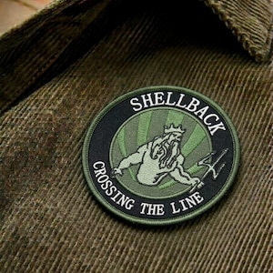 May include: A circular patch with the text "SHELLBACK CROSSING THE LINE" encircling a green illustration of a figure with a trident. The patch is black and green, and is sewn onto a textured, olive green fabric.