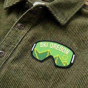 May include: Olive green corduroy shirt with a button-down collar and two front buttons. A green patch with the words "SKI OREGON" and a ski goggles design is sewn on the shirt.
