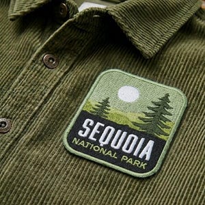 May include: Close-up of an olive green corduroy shirt with a "Sequoia National Park" patch. The patch features a landscape with trees, a moon, and the park's name in white lettering. The shirt has a button-down collar.