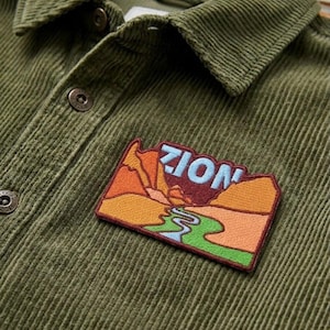 May include: Close-up of a green corduroy shirt with a "ZION" patch. The patch features a landscape design with mountains, a river, and the word "ZION" in blue letters. The shirt has a button-down collar and two visible buttons.