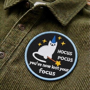 May include: Olive green corduroy shirt with a round patch featuring a white cat wearing a blue wizard hat and holding a wand. The patch reads "Hocus Pocus, you've now lost your focus" with yellow star accents.