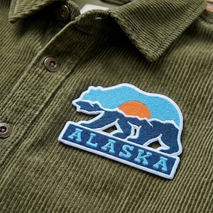 May include: Olive green corduroy shirt with a button-down collar and a patch featuring a bear silhouette with a sunset design and the word "ALASKA" in blue and white.