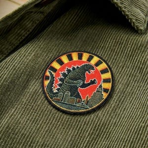 May include: Olive green corduroy shirt with a circular patch featuring a Godzilla design. The patch shows Godzilla in black, a red sun, and a cityscape. The shirt has a button-down collar and a textured corduroy fabric.