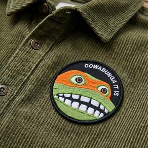 May include: Olive green corduroy shirt with a button-down collar and a round patch featuring a cartoon turtle face with the text "COWABUNGA IT IS". The shirt has brown buttons.