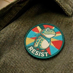 May include: Embroidered patch featuring a cartoon frog wearing a blue bandana, set against a colorful starburst design. The word "RESIST" is embroidered in white. The patch is sewn onto a green corduroy fabric.