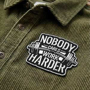 May include: Close-up of an olive green corduroy shirt with a black and white patch. The patch reads "NOBODY CARES WORK HARDER" with a barbell graphic. The shirt has a button-down collar and two visible buttons.