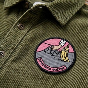 May include: Close-up of an olive green corduroy shirt with a round patch. The patch features a hand sweeping with a broom, with the words "problem solver" in a pink and gray circle.