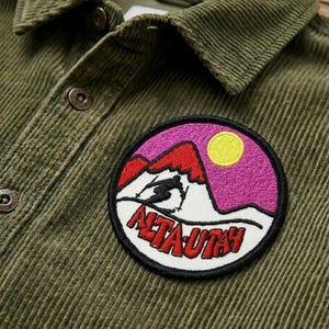 May include: Olive green corduroy shirt with a circular embroidered patch. The patch features a skier on a snowy mountain with the text "ALTA UTAH" in red. The background of the patch is pink and yellow.