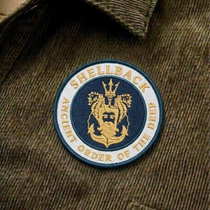 May include: A circular patch with the text "SHELLBACK ANCIENT ORDER OF THE DEEP" in white lettering. The patch features a navy blue center with a gold emblem of a trident, seahorses, and an anchor. The patch is sewn onto a green corduroy fabric.