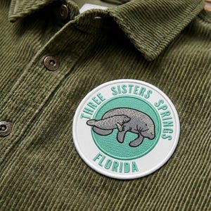 May include: Olive green corduroy shirt with a round patch featuring a manatee and the text "THREE SISTERS SPRINGS FLORIDA" in a circular design. The shirt has a button-down collar and two front buttons.