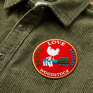 May include: Olive green corduroy shirt with a button-down collar. A circular patch with the words "LOVE, PEACE, MUSIC, WOODSTOCK" surrounds a white dove perched on a guitar. The patch is red with yellow trim.