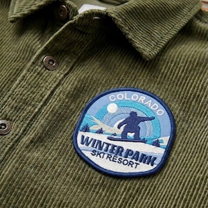 May include: Close-up of an olive green corduroy shirt with a button-down collar. A circular patch on the shirt features a skier silhouette, the text "Colorado", and "Winter Park Ski Resort".