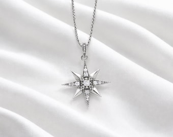 May include: A silver-toned necklace with a starburst pendant. The pendant features a central round gemstone surrounded by smaller gemstones and pointed rays. The necklace hangs on a delicate chain against a white fabric background.