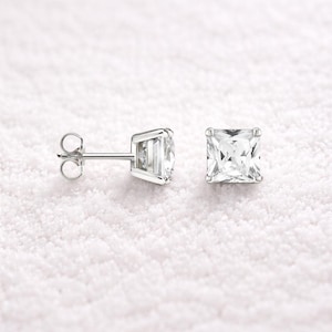May include: A pair of square-cut, clear gemstone stud earrings. The earrings are set in a silver-colored metal and are displayed on a white, textured surface. The earrings have a classic, elegant design.