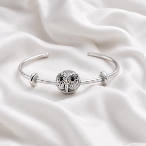 May include: A silver-toned cuff bracelet featuring a central owl charm with detailed feather engravings and two black circular eyes. The bracelet includes two small silver-toned spacer beads positioned on either side of the owl charm.