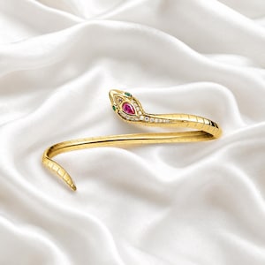 May include: A gold-toned snake bracelet featuring a textured, segmented body. The snake head is embellished with a central pear-shaped pink stone, small clear accent stones along the snout, and two small green stones serving as the eyes.