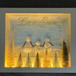 Pode incluir: Uma cena de inverno emoldurada com as palavras douradas "Let it Snow". A obra apresenta três bonecos de neve de mãos dadas, com um fundo de paisagem nevada e árvores de Natal em miniatura iluminadas. A moldura é branca.