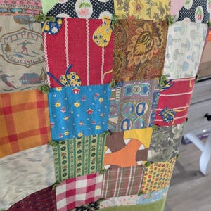 Może przedstawiać: Narzuta typu patchwork składająca się z różnych kwadratów materiału o zróżnicowanych wzorach, w tym kwiatowych, w kratę, vichy, paisley oraz z grafiką narciarza. Kwadraty są połączone małymi zielonymi węzełkami z tkaniny na rogach.