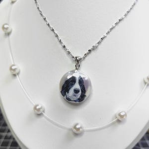 Puede incluir: Un collar de plata con un colgante que presenta un retrato de perro en blanco y negro. El colgante redondo cuelga de una delicada cadena de plata. También es visible un collar separado con perlas blancas.