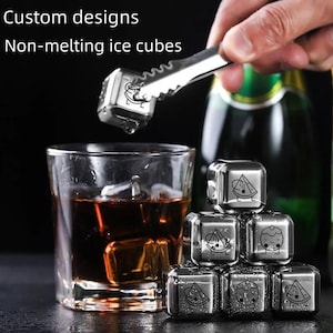 May include: Stainless steel non-melting ice cubes with custom designs, held by tongs, next to a glass of amber liquid. The cubes have various food-themed illustrations. Text reads "Custom designs" and "Non-melting ice cubes."