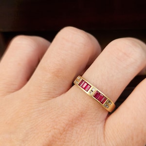 May include: A gold band ring featuring a row of rectangular, deep red rubies set horizontally. Small, clear, round-cut diamonds are interspersed between the rubies and placed at the ends of the stone setting, creating a linear, multi-stone design.
