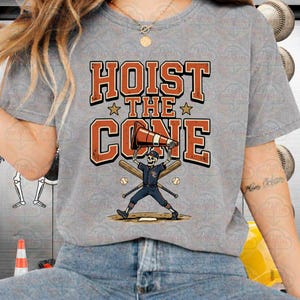 May include: Gray t-shirt with the text "HOIST THE CONE" in orange and white. The graphic features a skeleton baseball player holding a traffic cone, with baseball bats and balls. A fun, sporty design.
