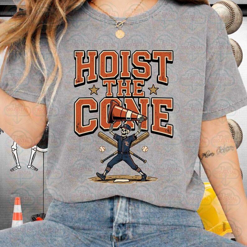 May include: Gray t-shirt with the text "HOIST THE CONE" in orange and white. The graphic features a skeleton baseball player holding a traffic cone, with baseball bats and balls. A fun, sporty design.