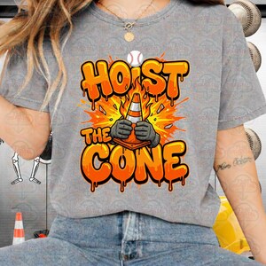 May include: A gray t-shirt with the words "HOIST THE CONE" in orange, featuring a baseball and a traffic cone graphic. The cone is held by gloved hands, with an explosion effect in the background.