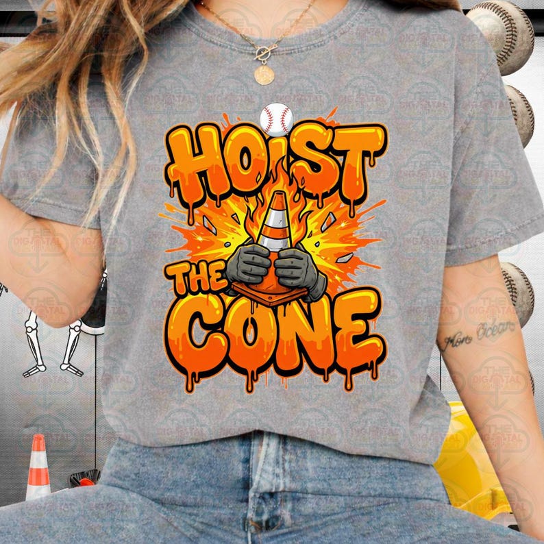 May include: A gray t-shirt with the words "HOIST THE CONE" in orange, featuring a baseball and a traffic cone graphic. The cone is held by gloved hands, with an explosion effect in the background.