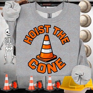 以下が含まれることがあります： オレンジ色のブロック文字で「HOIST THE CONE」と書かれたグレーのスウェットシャツ。オレンジと白のトラフィックコーンのグラフィックが特徴です。野球と建設をテーマにしたアイテムが背景にあります。