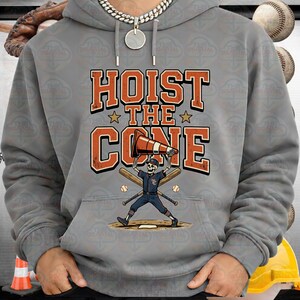 May include: Gray hoodie with a graphic that says "HOIST THE CONE". The graphic features a skeleton baseball player holding a traffic cone, with baseball bats and balls. The hoodie has a drawstring hood.