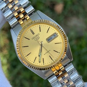 May include: A gold-toned Seiko 5 automatic watch with a gold dial and a two-tone silver and gold stainless steel band. The watch face displays the day and date. The watch is new and still has protective plastic on the band.
