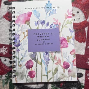 May include: A spiral-bound journal with a floral design in pink, purple, and blue. The cover reads "Bloom Where You Are Planted" and features a purple label with the text "Proverbs 31 Woman Journal by Barbara Hubert."