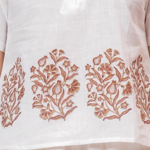 May include: White linen top with short sleeves and a round neckline. The top has pearl-like buttons down the front and a floral pattern in brown and tan across the lower section. The floral design includes stylized flowers and leaves.