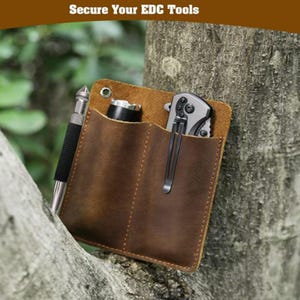 Puede incluir: Un organizador EDC (Everyday Carry) de cuero marrón con tres bolsillos, que contiene un bolígrafo, una linterna y una navaja plegable. El organizador está sujeto a un tronco de árbol. El texto "Secure Your EDC Tools" está en la parte superior.
