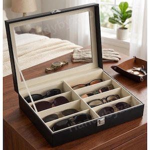 May include: A black sunglasses organizer with a mirrored lid, showcasing various sunglasses in individual compartments. The interior is lined with a light beige fabric. A watch and gloves are reflected in the mirror.