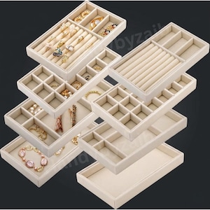 May include: Beige velvet jewelry organizer trays. The trays have various compartments for rings, bracelets, necklaces, and other jewelry. The trays are stacked, showcasing the different storage options. The background is a dark gray.