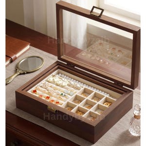 May include: A wooden jewelry box with a clear glass lid, open to reveal various gold and pearl jewelry pieces. The box is rectangular with a dark brown finish and a beige interior. A small mirror and a book are visible.