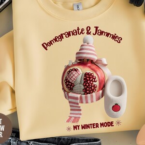 May include: Light yellow sweatshirt with a whimsical illustration of a pomegranate wearing a hat, scarf, and slippers. Text reads "Pomegranate & Jammies" and "My Winter Mode".
