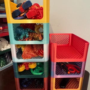 Op de afbeelding: Stapelbare, kleurrijke plastic opbergbakken in geel, turquoise, wit, groen en paars. De bakken zijn gevuld met diverse items, waaronder speelgoed en knutselspullen. De verticale opstelling van de bakken biedt een ruimtebesparende opbergoplossing.