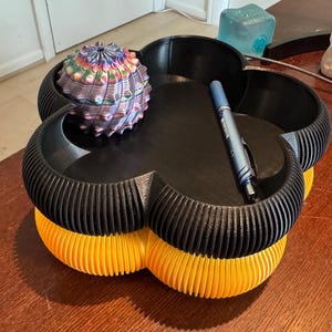 May include: A unique, multi-colored decorative object resembling a cactus sits on a black and yellow flower-shaped tray. A blue pen rests in one of the tray's compartments. The tray has a ribbed design and is made of plastic.