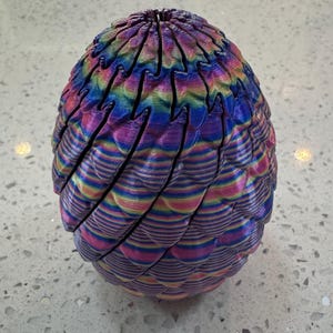 May include: A colorful, decorative egg-shaped object with a textured, layered design. The egg features a vibrant spectrum of colors, including pink, blue, yellow, and purple, arranged in horizontal stripes. The top of the egg has a unique, open design.