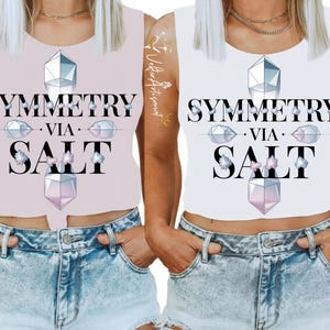 Peut inclure: Deux débardeurs sans manches, un rose clair et un blanc, chacun avec le texte "SYMMETRY VIA SALT" en noir. Le design comprend des formes de cristaux géométriques et des accents floraux. Les deux hauts sont associés à des shorts en jean.