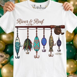 Peut inclure: T-shirt blanc avec un motif de canne à pêche et divers leurres suspendus en dessous. Le texte "River & Reef *1776*" est imprimé au-dessus. Le t-shirt est tenu par une personne.