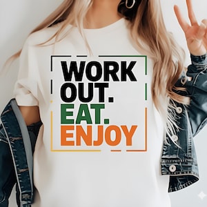 Peut inclure: Un t-shirt crème avec les mots "WORK OUT. EAT. ENJOY" en noir, vert et orange. Le texte est encadré de lignes noires, vertes et orange. Le t-shirt est un vêtement décontracté.